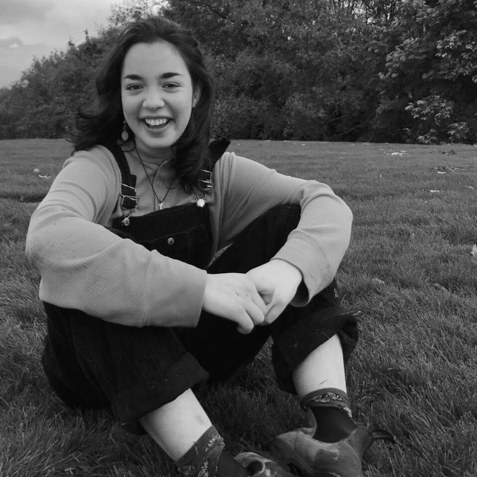 Maria sits in a park mid-laugh. She has dark brown shoulder length hair and is wearing overalls and an off-white T-shirt.